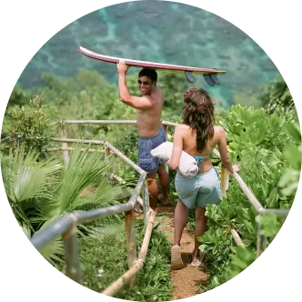 Man holding a surfboard on his head with a woman following walking on a boardwalk among exotic plants